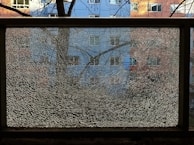 A shattered glass pane is framed by a black metal border, reflecting a fragmented image of a residential building with blue and pink sections. Bare tree branches are visible against the window, creating a textured overlay.