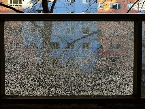 A shattered glass pane is framed by a black metal border, reflecting a fragmented image of a residential building with blue and pink sections. Bare tree branches are visible against the window, creating a textured overlay.