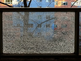 A shattered glass pane is framed by a black metal border, reflecting a fragmented image of a residential building with blue and pink sections. Bare tree branches are visible against the window, creating a textured overlay.
