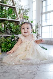 A baby in a delicate, flower-embroidered dress sits happily on a patterned floor. The setting includes lush green plants and flowers near a wooden ladder, with soft natural light streaming in through large windows.