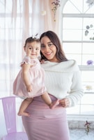 A woman is holding a baby, both smiling and looking at the camera. The woman is wearing a white sweater and a pink skirt, while the baby is dressed in a pink dress. The background includes a window with sheer curtains and hanging flowers, creating a soft, delicate atmosphere.