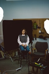 A person is seated on a stool in what appears to be a photography studio or workshop setting. The individual is wearing a gray sweater over a white shirt and blue jeans. Behind the person, there is a black backdrop hanging on a pegboard wall with various tools and items, such as a green backpack and some boxing gloves. The room is lit with studio lights, and there is an empty chair nearby, along with other studio equipment like tripods and cables.