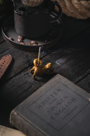 A dimly lit scene featuring a vintage book with the title 'Written and Spoken English', placed on a dark wooden surface. Nearby, a small, sculpted incense holder in the shape of a cat emits a thin trail of smoke. In the background, there is a rustic metal coffee pot with coffee beans scattered around.