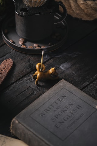 A dimly lit scene featuring a vintage book with the title 'Written and Spoken English', placed on a dark wooden surface. Nearby, a small, sculpted incense holder in the shape of a cat emits a thin trail of smoke. In the background, there is a rustic metal coffee pot with coffee beans scattered around.
