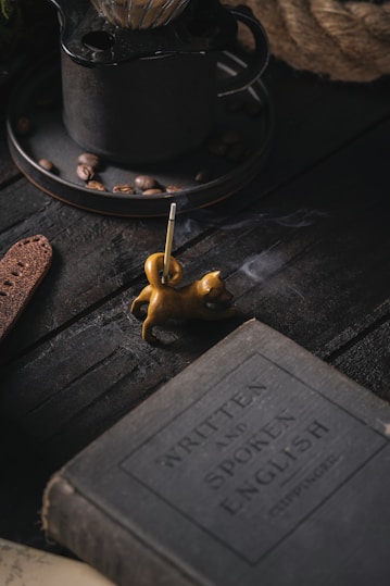 A dimly lit scene featuring a vintage book with the title 'Written and Spoken English', placed on a dark wooden surface. Nearby, a small, sculpted incense holder in the shape of a cat emits a thin trail of smoke. In the background, there is a rustic metal coffee pot with coffee beans scattered around.