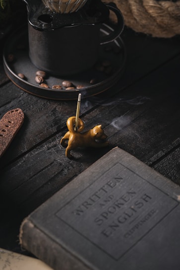 A dimly lit scene featuring a vintage book with the title 'Written and Spoken English', placed on a dark wooden surface. Nearby, a small, sculpted incense holder in the shape of a cat emits a thin trail of smoke. In the background, there is a rustic metal coffee pot with coffee beans scattered around.