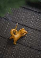 A small, wooden cat-shaped pencil holder with a pencil inserted into it is placed on a dark, textured mat. The cat is in a stretching pose with its back arched.