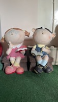 Two large, playful figurines of children sitting on benches. One figure is a girl with pigtails wearing a pink dress and yellow socks, holding a red book. The other is a boy in overalls and a cap, also reading a book. Both have exaggerated round faces with smiling expressions and appear in a whimsical style.