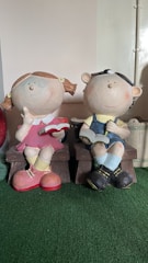 Two large, playful figurines of children sitting on benches. One figure is a girl with pigtails wearing a pink dress and yellow socks, holding a red book. The other is a boy in overalls and a cap, also reading a book. Both have exaggerated round faces with smiling expressions and appear in a whimsical style.