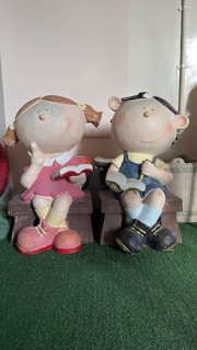 Two large, playful figurines of children sitting on benches. One figure is a girl with pigtails wearing a pink dress and yellow socks, holding a red book. The other is a boy in overalls and a cap, also reading a book. Both have exaggerated round faces with smiling expressions and appear in a whimsical style.