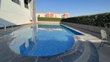 A serene outdoor swimming pool with clear blue water is surrounded by a stone deck. One side features a line of manicured hedges, and a white building is partially visible on the left. A metal pool ladder is present in the foreground, while a single chair sits on the right side of the deck.