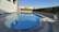 A serene outdoor swimming pool with clear blue water is surrounded by a stone deck. One side features a line of manicured hedges, and a white building is partially visible on the left. A metal pool ladder is present in the foreground, while a single chair sits on the right side of the deck.