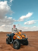A person wearing a headscarf is seated on a yellow all-terrain vehicle (ATV) on a vast desert landscape under a blue sky with scattered clouds.