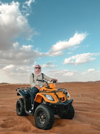 A person wearing a headscarf is seated on a yellow all-terrain vehicle (ATV) on a vast desert landscape under a blue sky with scattered clouds.