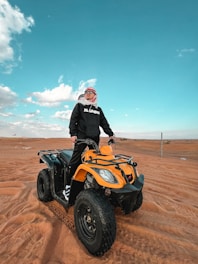 A person stands confidently on a yellow ATV in the middle of a vast desert landscape. The person is wearing a black hoodie and sunglasses, with a red and white headscarf. The sky is bright blue with scattered clouds, adding a vivid contrast to the sandy terrain.