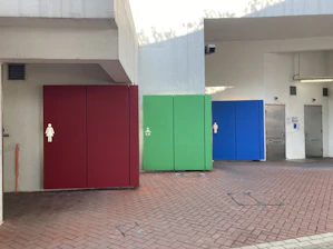 A public restroom area with distinct colored doors representing different facilities. The red door indicates a women's restroom, the green door seems to denote family or accessible facilities, and the blue door is marked for men. The setting appears to be constructed from concrete with a red brick pattern on the ground.