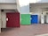 A public restroom area with distinct colored doors representing different facilities. The red door indicates a women's restroom, the green door seems to denote family or accessible facilities, and the blue door is marked for men. The setting appears to be constructed from concrete with a red brick pattern on the ground.