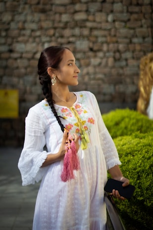 A woman with a long braid wears a white dress adorned with colorful floral embroidery. She holds a tassel and a phone, standing against a background of a stone wall and green bushes.