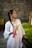 A woman with a long braid wears a white dress adorned with colorful floral embroidery. She holds a tassel and a phone, standing against a background of a stone wall and green bushes.