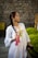 A woman with a long braid wears a white dress adorned with colorful floral embroidery. She holds a tassel and a phone, standing against a background of a stone wall and green bushes.