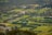 Aerial view of a lush, rural landscape with green fields and patches of trees. Various houses and farm buildings are scattered throughout the area, with roads connecting different sections. The landscape is divided into well-defined parcels of land, with a mix of open grass fields and wooded areas. In the distance, a small body of water is visible, surrounded by trees.
