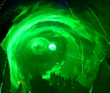 A group of silhouetted people are standing before a large, swirling green vortex or portal that is illuminated with bright light. The green lighting creates a mystical and otherworldly atmosphere.