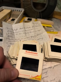 A collection of vintage Kodachrome slide transparencies lies on a table, surrounded by handwritten letters on lined paper. Some slides are stacked, while one nearest the camera is held in hand. In the background, a circular slide holder and more boxes of slides can be seen.