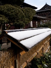 A traditional wooden fence topped with a thin layer of snow, set against a background of well-maintained, intricately pruned trees and a traditional building with ornate wooden architecture.
