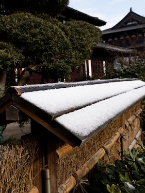 A traditional wooden fence topped with a thin layer of snow, set against a background of well-maintained, intricately pruned trees and a traditional building with ornate wooden architecture.