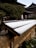A traditional wooden fence topped with a thin layer of snow, set against a background of well-maintained, intricately pruned trees and a traditional building with ornate wooden architecture.
