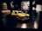 A model car with a glossy yellow finish and black stripes is placed on a dark surface. The car doors are open, revealing some interior details. The background is blurred and dark, creating a dramatic contrast with the vibrant model car.