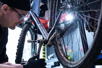 A person wearing a hat and glasses is repairing a bicycle in a workshop. The focus is on the drivetrain area, with a wrench being used to adjust a component. The setting is industrial with tools and equipment visible in the background.