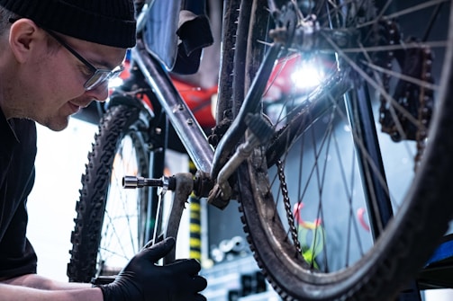 A person wearing a hat and glasses is repairing a bicycle in a workshop. The focus is on the drivetrain area, with a wrench being used to adjust a component. The setting is industrial with tools and equipment visible in the background.