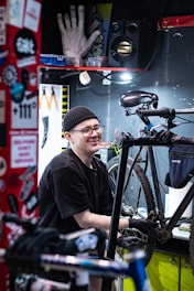 A person wearing a black beanie and glasses is working on a bicycle in a well-organized workshop. Various tools are visible, including pliers hanging on a pegboard. The workspace is adorned with stickers and colorful decorations.