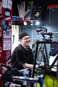 A person wearing a black beanie and glasses is working on a bicycle in a well-organized workshop. Various tools are visible, including pliers hanging on a pegboard. The workspace is adorned with stickers and colorful decorations.