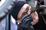 A person, wearing glasses and a black beanie, is intently inspecting a bicycle frame. They are using a tool to work on the bike and are wearing black gloves. The close-up image focuses on their concentrated expression and the metalwork of the bike.
