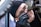A person, wearing glasses and a black beanie, is intently inspecting a bicycle frame. They are using a tool to work on the bike and are wearing black gloves. The close-up image focuses on their concentrated expression and the metalwork of the bike.