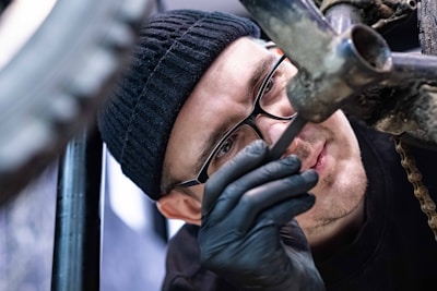 A person, wearing glasses and a black beanie, is intently inspecting a bicycle frame. They are using a tool to work on the bike and are wearing black gloves. The close-up image focuses on their concentrated expression and the metalwork of the bike.