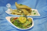 The image features a plate of golden-brown battered fish fillets sitting atop a pile of fries. Next to it is a fresh salad with a variety of vegetables, including lettuce, cherry tomatoes, cucumber, and a rolled slice of lemon as a garnish, all arranged on a white plate. A small bowl of green herbs or sauce accompanies the dish, and there is a white wrapped item tied with a string. The background is a soft blue fabric.