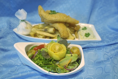 The image features a plate of golden-brown battered fish fillets sitting atop a pile of fries. Next to it is a fresh salad with a variety of vegetables, including lettuce, cherry tomatoes, cucumber, and a rolled slice of lemon as a garnish, all arranged on a white plate. A small bowl of green herbs or sauce accompanies the dish, and there is a white wrapped item tied with a string. The background is a soft blue fabric.