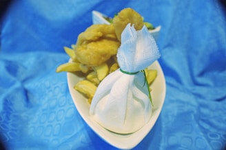 A plate of fried food, including chips and presumably fish or chicken nuggets, sits on a blue tablecloth. A small wrapped item is tied with a green string, possibly containing a condiment or lemon for squeezing.