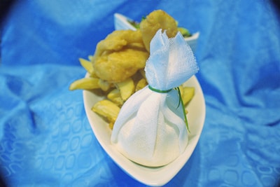 A plate of fried food, including chips and presumably fish or chicken nuggets, sits on a blue tablecloth. A small wrapped item is tied with a green string, possibly containing a condiment or lemon for squeezing.