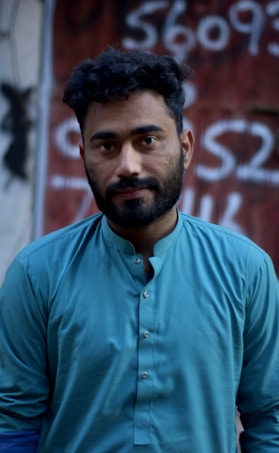A man with short curly hair and a beard, wearing a turquoise shirt, stands in front of a blurred background featuring graffiti-like numbers and letters on a dark red surface.