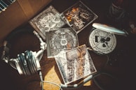A collection of metal music CDs is spread out on a wooden surface. The artwork on the CD covers features intricate, dark designs with skulls and abstract patterns. A pair of silver and black headphones lie next to a portable CD player.