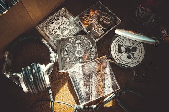 A collection of metal music CDs is spread out on a wooden surface. The artwork on the CD covers features intricate, dark designs with skulls and abstract patterns. A pair of silver and black headphones lie next to a portable CD player.