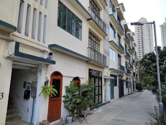 A row of mid-rise apartment buildings with balconies and large windows, exhibiting a somewhat vintage architectural style. The ground floor has several doors, some surrounded by potted plants, creating a homely atmosphere. The scene includes a quiet sidewalk lined with trees and a modern streetlight.