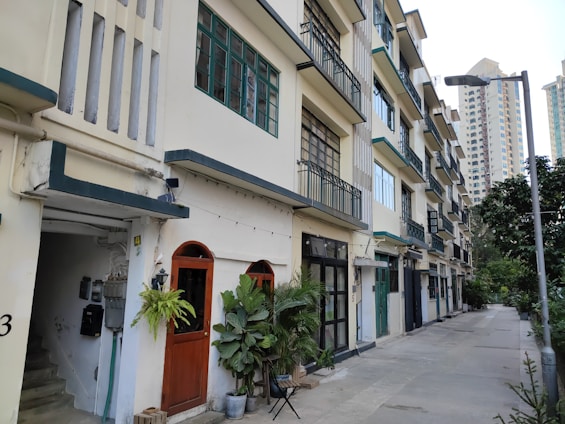 A row of mid-rise apartment buildings with balconies and large windows, exhibiting a somewhat vintage architectural style. The ground floor has several doors, some surrounded by potted plants, creating a homely atmosphere. The scene includes a quiet sidewalk lined with trees and a modern streetlight.