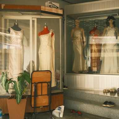 A storefront display featuring several mannequins wearing elegant dresses. The setting appears to be a vintage or retro shop with muted lighting. In front of the display, there is a folded wooden table and a potted plant. Various small items, including shoes, are scattered on the ground.