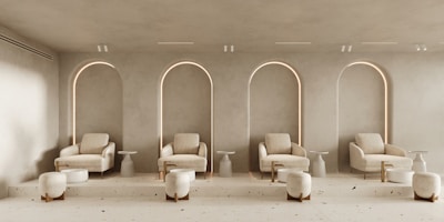 A modern, minimalist interior featuring a row of four cream-colored armchairs, each paired with a small round side table and a short cylindrical ottoman. The chairs are set against a wall with arched recesses, softly lit from behind, creating a warm ambiance. The floor is light-colored with subtle speckles, complementing the serene, neutral color palette of the room.