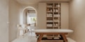 A minimalist, serene spa room features a massage table with wooden legs and a white cushioned surface. To the right, a wall-mounted cabinet with open shelves holds neatly organized spa products, including bottles and jars. In the background, an archway leads to another area with a simple chair and a countertop that holds decorative items. Soft, neutral tones dominate the space, creating a tranquil atmosphere.